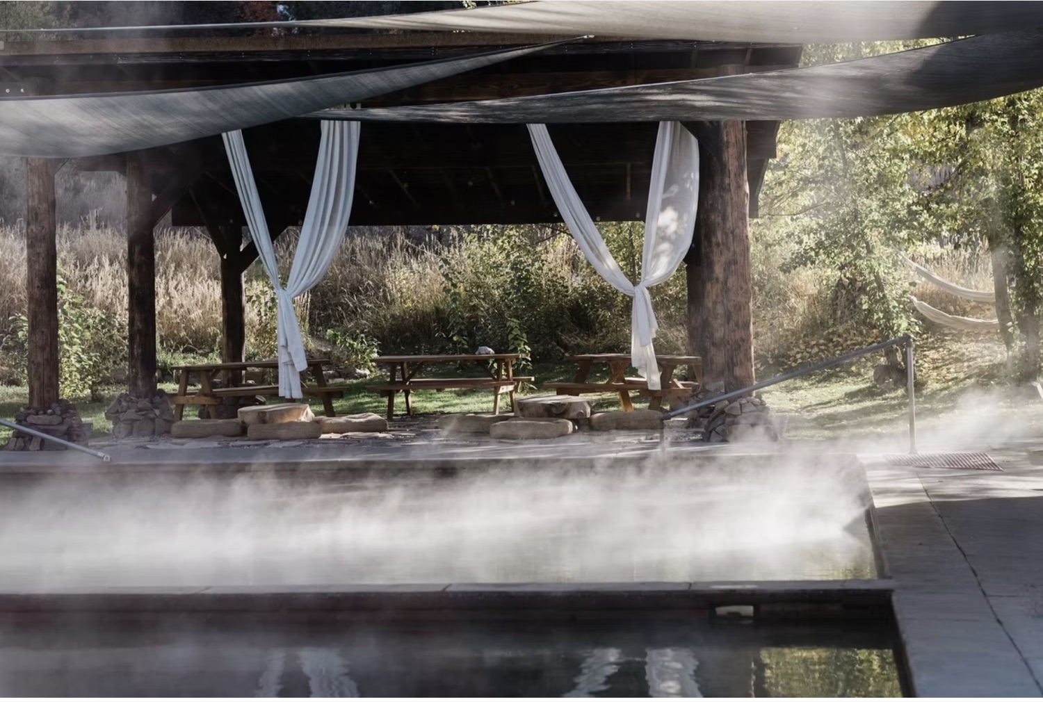 Steam rising off a hot pool beside a covered pavilion with white curtains